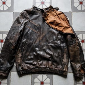 Puede incluir: Chaqueta de cuero marrón desgastado con cuello alto y múltiples bolsillos con cremallera. La chaqueta tiene un aspecto desgastado y vintage con un forro marrón en contraste visible en el cuello. La chaqueta está tendida sobre una superficie de baldosas estampadas.