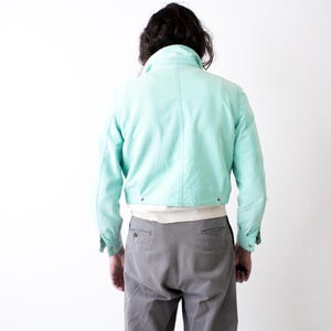 May include: A cropped, light turquoise jacket with a high collar and buttoned cuffs. The jacket is worn over a white shirt and paired with gray trousers. The model is viewed from the back against a white background.