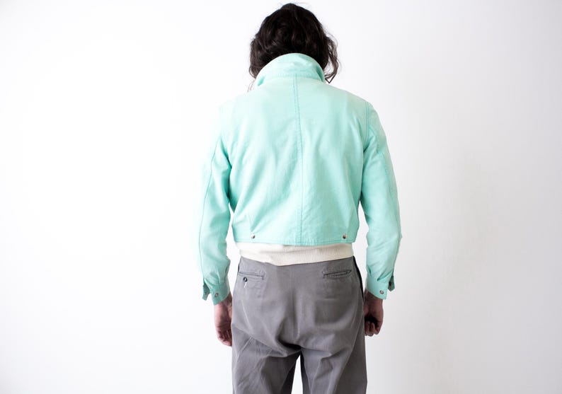 May include: A cropped, light turquoise jacket with a high collar and buttoned cuffs. The jacket is worn over a white shirt and paired with gray trousers. The model is viewed from the back against a white background.