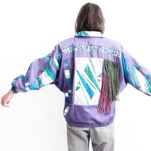 Könnte beinhalten: Eine lila Jacke mit Patchwork-Design in verschiedenen Farben und Mustern, darunter Grün, Blau und Weiß. Die Jacke hat einen Fransenbesatz auf der Rückseite.