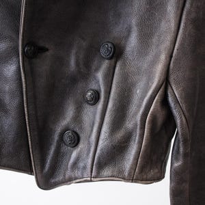 K&ouml;nnte beinhalten: Eine dunkelbraune Lederjacke mit zweireihigem Verschluss und vier sichtbaren schwarzen Kn&ouml;pfen. Das Leder hat eine texturierte, matte Oberfl&auml;che mit subtilen Farbnuancen, und das Kleidungsst&uuml;ck ist mit strukturierten N&auml;hten versehen.