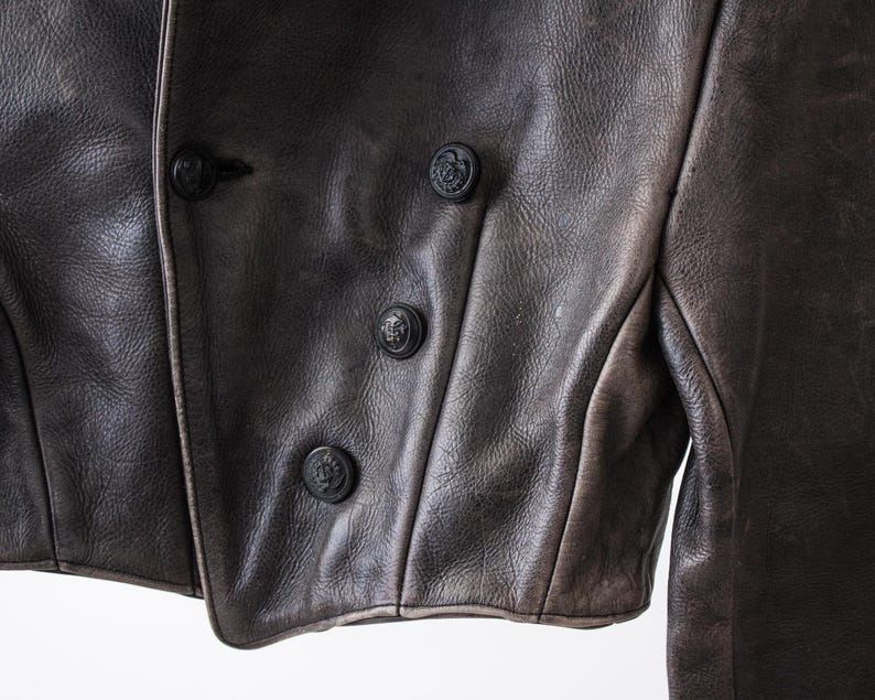 K&ouml;nnte beinhalten: Eine dunkelbraune Lederjacke mit zweireihigem Verschluss und vier sichtbaren schwarzen Kn&ouml;pfen. Das Leder hat eine texturierte, matte Oberfl&auml;che mit subtilen Farbnuancen, und das Kleidungsst&uuml;ck ist mit strukturierten N&auml;hten versehen.