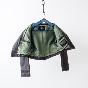 K&ouml;nnte beinhalten: Eine dunkelbraune Lederjacke mit hellgr&uuml;nem Innenfutter, die an einem blauen Holzkleiderb&uuml;gel mit der Aufschrift Bayard h&auml;ngt. Die Jacke verf&uuml;gt &uuml;ber zwei Innentaschen auf Brusth&ouml;he und ein eingen&auml;htes, rechteckiges schwarzes Markenetikett.