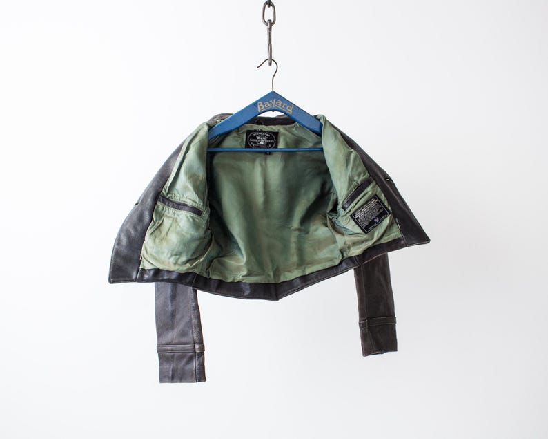 K&ouml;nnte beinhalten: Eine dunkelbraune Lederjacke mit hellgr&uuml;nem Innenfutter, die an einem blauen Holzkleiderb&uuml;gel mit der Aufschrift Bayard h&auml;ngt. Die Jacke verf&uuml;gt &uuml;ber zwei Innentaschen auf Brusth&ouml;he und ein eingen&auml;htes, rechteckiges schwarzes Markenetikett.