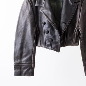 K&ouml;nnte beinhalten: Eine kurze, dunkelbraune Lederjacke mit breitem Reverskragen, langen &Auml;rmeln mit Manschetten und einer zweireihigen Knopfleiste mit drei runden, schwarzen Kn&ouml;pfen. Das gr&uuml;ne Innenfutter ist im Bereich des Kragens sichtbar.