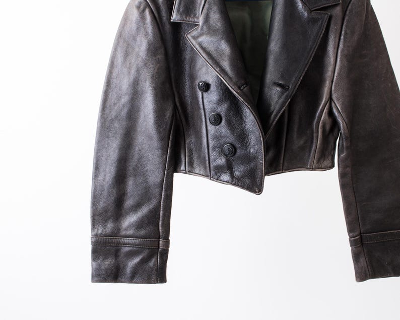 K&ouml;nnte beinhalten: Eine kurze, dunkelbraune Lederjacke mit breitem Reverskragen, langen &Auml;rmeln mit Manschetten und einer zweireihigen Knopfleiste mit drei runden, schwarzen Kn&ouml;pfen. Das gr&uuml;ne Innenfutter ist im Bereich des Kragens sichtbar.