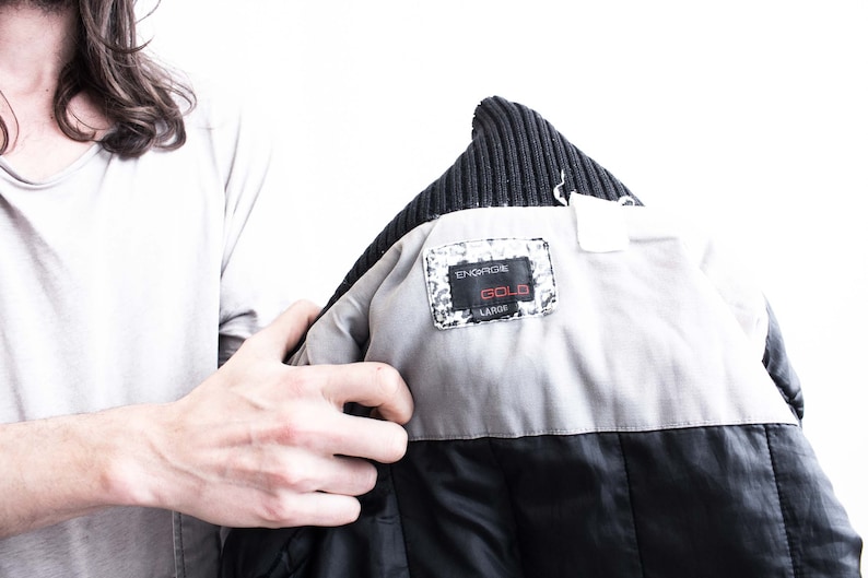 K&ouml;nnte beinhalten: Eine schwarze und graue Jacke mit einem schwarzen Strickkragen. Die Jacke hat ein wei&szlig;es Futter mit einem schwarzen und wei&szlig;en Etikett mit der Aufschrift "ENGORGON GOLD LARGE".