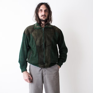 Op de afbeelding: Een vintage groene en bruine jas met een ritssluiting. De jas heeft een combinatie van gebreide en suède-achtige materialen. Het model draagt een grijze broek. De achtergrond is effen wit.