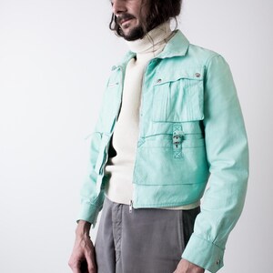 May include: A light turquoise jacket with multiple pockets and a zipper closure. The jacket is worn over a cream-colored turtleneck sweater and paired with gray pants. The jacket features a collared neckline and buttoned cuffs.