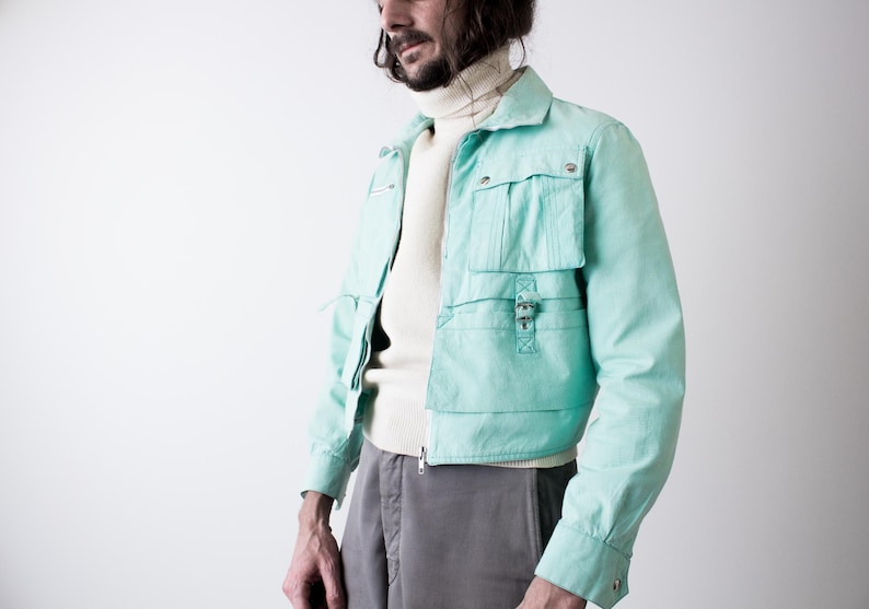 May include: A light turquoise jacket with multiple pockets and a zipper closure. The jacket is worn over a cream-colored turtleneck sweater and paired with gray pants. The jacket features a collared neckline and buttoned cuffs.
