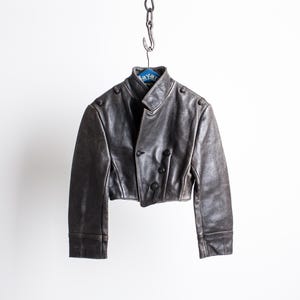 K&ouml;nnte beinhalten: Eine kurze, dunkelbraune Lederjacke mit Stehkragen, langen &Auml;rmeln mit Manschetten und einer zweireihigen Vorderseite mit drei sichtbaren dunklen Kn&ouml;pfen. Die Jacke h&auml;ngt an einem Kettenhaken auf einem blauen Kleiderb&uuml;gel.