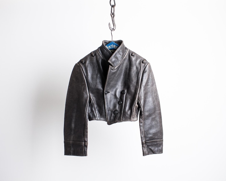 K&ouml;nnte beinhalten: Eine kurze, dunkelbraune Lederjacke mit Stehkragen, langen &Auml;rmeln mit Manschetten und einer zweireihigen Vorderseite mit drei sichtbaren dunklen Kn&ouml;pfen. Die Jacke h&auml;ngt an einem Kettenhaken auf einem blauen Kleiderb&uuml;gel.