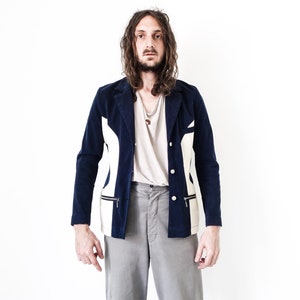 Puede incluir: Un hombre con una chaqueta azul marino y blanca con un detalle de cremallera. Lleva pantalones grises y tiene el pelo largo y casta&ntilde;o.
