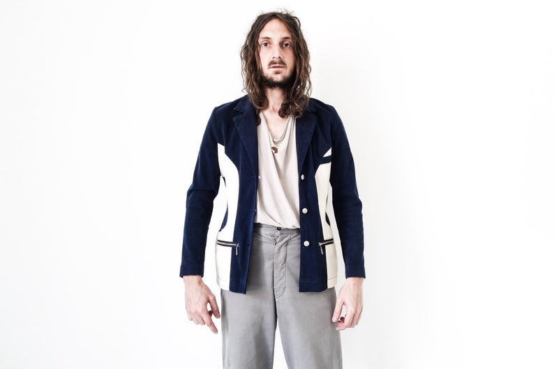 Puede incluir: Un hombre con una chaqueta azul marino y blanca con un detalle de cremallera. Lleva pantalones grises y tiene el pelo largo y casta&ntilde;o.