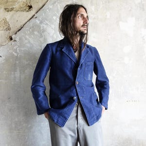 Può includere: Un blazer blu scuro con un solo bottone, due tasche anteriori e revers a lancia. Il blazer è indossato sopra una camicia di colore chiaro e abbinato a pantaloni grigi. Lo sfondo è un muro strutturato bianco sporco.