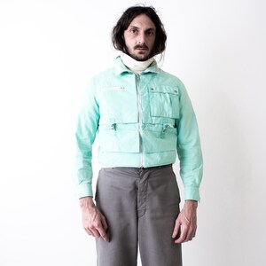 May include: A light turquoise jacket with multiple front pockets and a zip closure. The jacket is paired with gray trousers. The model is wearing a white turtleneck underneath the jacket.