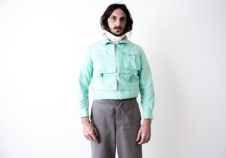 May include: A light turquoise jacket with multiple front pockets and a zip closure. The jacket is paired with gray trousers. The model is wearing a white turtleneck underneath the jacket.