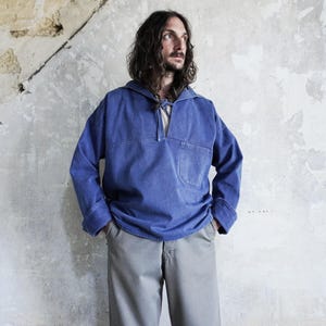 Pode incluir: Uma camisa azul de manga comprida com gola estilo marinheiro, fecho de cordão no decote e um bolso único no peito. A peça tem um corte largo com punhos amplos e é combinada com calças de cor cinzento-claro.