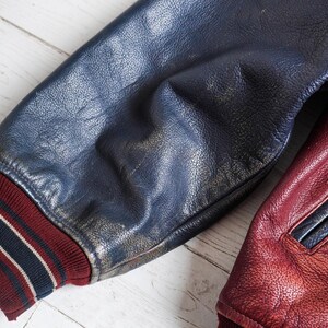 May include: A varsity jacket sleeve made of dark blue textured leather with a ribbed knit cuff in maroon, navy, and white stripes. Beside it, a portion of a maroon leather jacket panel is visible, featuring a matching maroon leather pocket trim.
