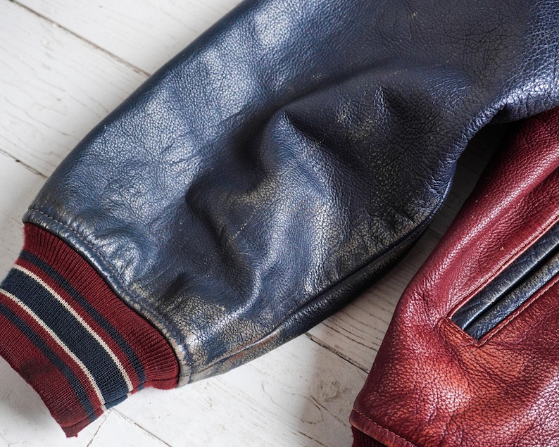 May include: A varsity jacket sleeve made of dark blue textured leather with a ribbed knit cuff in maroon, navy, and white stripes. Beside it, a portion of a maroon leather jacket panel is visible, featuring a matching maroon leather pocket trim.