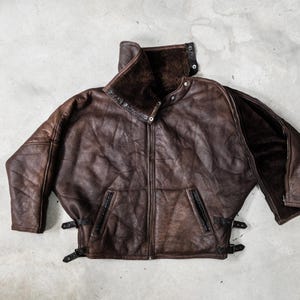 May include: A brown leather jacket with a high collar and a zip-up front. The jacket has two front pockets with zippers and adjustable straps at the waist. The interior lining is a darker brown, and the jacket appears to be vintage.