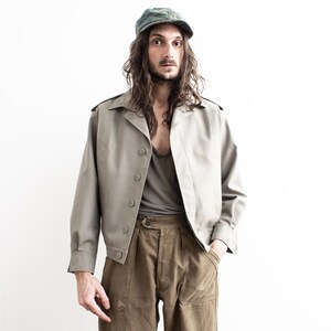 Pu&ograve; includere: Giacca verde oliva chiaro con cappellino abbinato. La giacca ha una chiusura con bottoni, spalline e maniche lunghe. Il modello indossa una canotta grigia e pantaloni verde oliva.