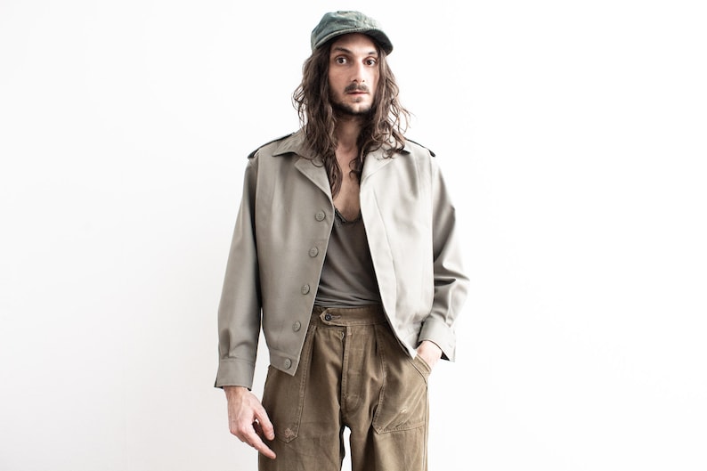Pu&ograve; includere: Giacca verde oliva chiaro con cappellino abbinato. La giacca ha una chiusura con bottoni, spalline e maniche lunghe. Il modello indossa una canotta grigia e pantaloni verde oliva.
