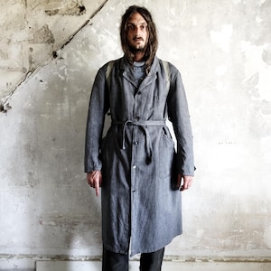May include: A gray, long coat with a matching belt. The coat has a button-up front and pockets. The person is wearing a light blue sweater underneath. The background is a distressed, textured wall.