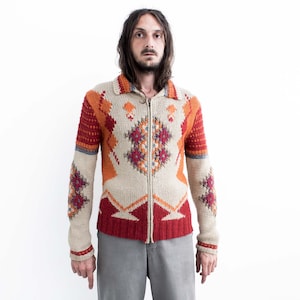 Könnte beinhalten: Ein beigefarbener, orangefarbener und roter Reißverschluss-Cardigan mit geometrischen Mustern. Der Cardigan hat einen Kragen und lange Ärmel. Das Design zeigt Rauten- und Zickzackformen in Orange-, Rot- und Beigetönen. Der Cardigan wird über einem grauen Hemd getragen.