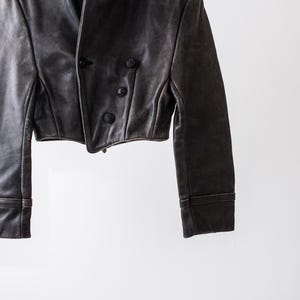 K&ouml;nnte beinhalten: Eine kurze, dunkelbraune Lederjacke mit zweireihigem Verschluss, drei sichtbaren runden Kn&ouml;pfen, langen &Auml;rmeln mit abgesteppten B&uuml;ndchen und einem strukturierten Kragen. Das Kleidungsst&uuml;ck hat eine glatte Textur und eine taillierte Passform.
