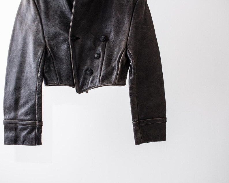 K&ouml;nnte beinhalten: Eine kurze, dunkelbraune Lederjacke mit zweireihigem Verschluss, drei sichtbaren runden Kn&ouml;pfen, langen &Auml;rmeln mit abgesteppten B&uuml;ndchen und einem strukturierten Kragen. Das Kleidungsst&uuml;ck hat eine glatte Textur und eine taillierte Passform.