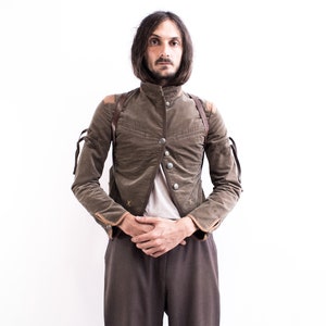May include: A man wearing a brown corduroy jacket with a high collar and leather straps. The jacket has a button closure and is worn over a white t-shirt. The man is standing with his hands clasped in front of him.