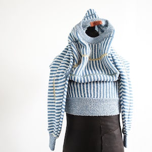 Op de afbeelding: Een blauw-wit gestreepte hoodie met lange mouwen. De trui heeft een textuur gebreid ontwerp en een lichtblauwe kleur. Een goudkleurige streep loopt over de voorkant. De trui wordt gepresenteerd op een zwarte vorm.