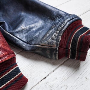 May include: A varsity jacket sleeve made of dark blue leather with a distressed finish, featuring a ribbed knit cuff in deep red with black and white stripes. A second sleeve in textured red leather with a matching striped cuff is visible.