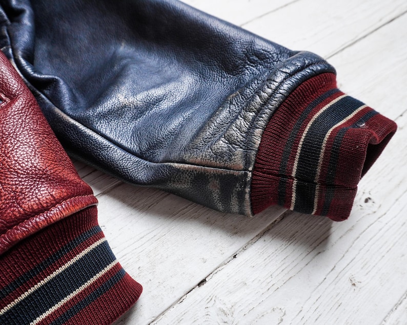 May include: A varsity jacket sleeve made of dark blue leather with a distressed finish, featuring a ribbed knit cuff in deep red with black and white stripes. A second sleeve in textured red leather with a matching striped cuff is visible.
