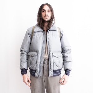 Puede incluir: Una chaqueta acolchada gris con cierre de cremallera y dos bolsillos grandes. La chaqueta tiene un cuello y puños de canalé.