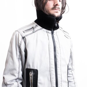K&ouml;nnte beinhalten: Eine wei&szlig;e Rei&szlig;verschlussjacke mit schwarzen N&auml;hten und einem schwarzen Stehkragen. Die Jacke hat einen Rei&szlig;verschluss und mehrere Taschen.
