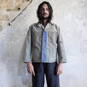 May include: A long-sleeved, light olive-green work jacket featuring a contrasting vertical blue fabric panel along the button placket, a standard collar, and two large patch pockets on the lower front, showing signs of wear and paint splatters.