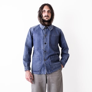 Op de afbeelding: Een blauw spijkerjack met een knoopsluiting, twee voorzakken en een ronde kraag. De jas wordt gedragen met een grijze broek. De algehele stijl is casual en geïnspireerd op werkkleding.