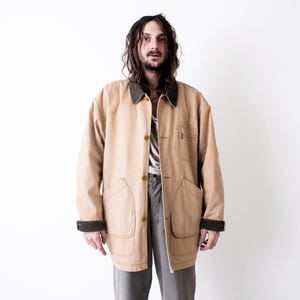 Può includere: Giacca da lavoro color cachi con colletto e polsini verde scuro. La giacca ha ampie tasche e chiusure con bottoni. La persona indossa una camicia a righe e pantaloni grigi. La giacca è uno stile workwear classico.