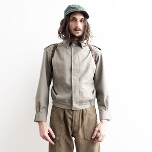 Pu&ograve; includere: Una persona indossa un berretto verde oliva vintage, una giacca verde oliva chiaro con spalline in pelle marrone e pantaloni marroni. La giacca ha un colletto button-down e maniche lunghe. I pantaloni hanno grandi tasche.