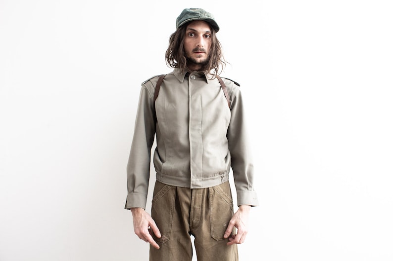Pu&ograve; includere: Una persona indossa un berretto verde oliva vintage, una giacca verde oliva chiaro con spalline in pelle marrone e pantaloni marroni. La giacca ha un colletto button-down e maniche lunghe. I pantaloni hanno grandi tasche.