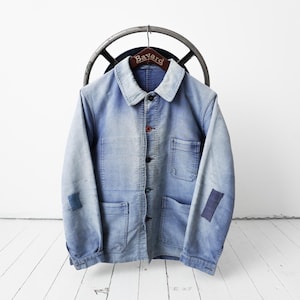 Pode incluir: Um casaco de trabalho azul-claro com acabamento desbotado, apresentando gola pontiaguda, fecho frontal com botões e três bolsos frontais. As mangas têm remendos retangulares de tecido azul-escuro e a peça está pendurada num cabide de madeira Bayard.