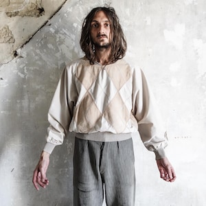 May include: A beige and tan argyle patterned sweater with long sleeves and a ribbed hem. The sweater is paired with grey, vertically striped trousers. The background is a textured, off-white wall.