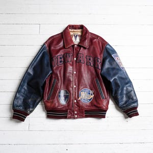 May include: A burgundy leather varsity jacket with navy blue sleeves, featuring the word NEWARK in dark blue lettering across the chest, multiple circular patches on the front, a shield patch on the left sleeve, and striped ribbed cuffs and hem.