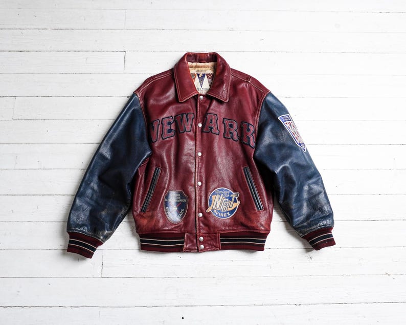 May include: A burgundy leather varsity jacket with navy blue sleeves, featuring the word NEWARK in dark blue lettering across the chest, multiple circular patches on the front, a shield patch on the left sleeve, and striped ribbed cuffs and hem.