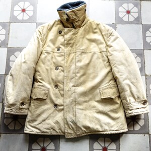 Könnte beinhalten: Eine cremefarbene, langärmelige Jacke mit hohem Kragen und Knopfleiste. Die Jacke hat zwei Taschen auf der Vorderseite und einen Reißverschluss. Die Jacke ist aus einem dicken, strapazierfähigen Stoff gefertigt und hat einen leicht abgenutzten Look.