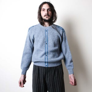 May include: A light blue cardigan with a ribbed texture, featuring a vertical blue stripe down the front and on the shoulders. The cardigan has a button-up closure and is paired with black and gray striped pants. The overall style is vintage.