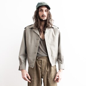 Pu&ograve; includere: Giacca color beige chiaro con bottoni e spalline. La giacca &egrave; abbinata a pantaloni color oliva e una maglietta grigia. Un berretto verde &egrave; indossato sulla testa.