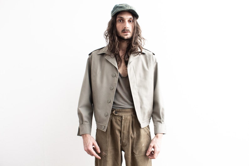 Pu&ograve; includere: Giacca color beige chiaro con bottoni e spalline. La giacca &egrave; abbinata a pantaloni color oliva e una maglietta grigia. Un berretto verde &egrave; indossato sulla testa.